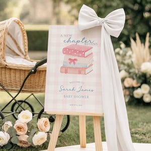 May include: A baby shower welcome sign on a wooden easel. The sign features a stack of watercolor books and the text "A new chapter begins" and "Welcome to Sarah James Baby Shower". A large white bow adorns the top.