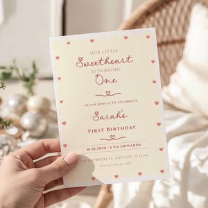 Könnte beinhalten: Eine cremefarbene Einladung mit Herzrand kündigt einen ersten Geburtstag an. Der Text lautet "Our Little Sweetheart is Turning One" und enthält den Namen "Sarah" und Veranstaltungsdetails. Die Einladung wird von einer Hand gehalten.
