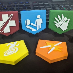 Puede incluir: Cinco fichas de plástico impresas en 3D con diferentes símbolos. Las fichas tienen forma de escudo con bordes redondeados. Las fichas son rojas, azules, verdes, amarillas y naranjas. Los símbolos son una cruz con una bala, una persona bajando las escaleras, una mano sosteniendo una barra, una persona corriendo en círculo y una estrella con dos líneas.