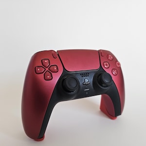 May include: A red and black video game controller with a matte finish. The controller features a directional pad, action buttons, and analog sticks. The PlayStation logo is centered on the controller.