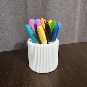May include: A white pen holder with a ribbed design, containing a selection of coloured markers. The markers have caps in shades such as blue, pink, yellow, and green. The holder is on a dark wooden surface.