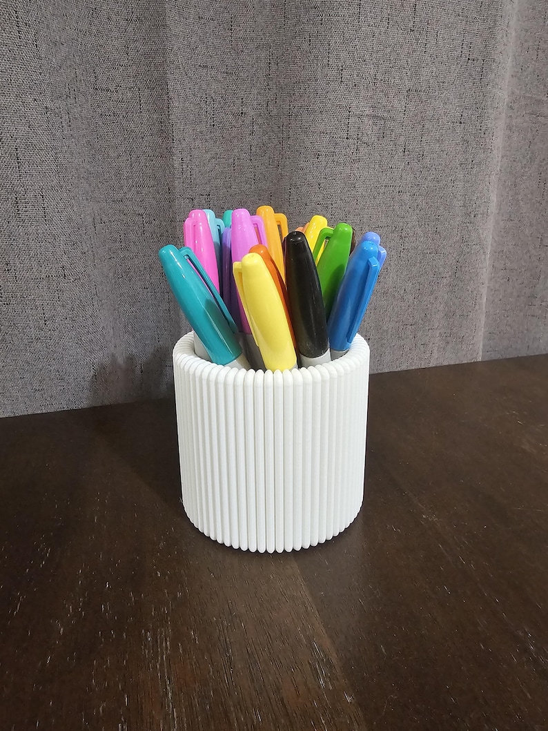 May include: A white pen holder with a ribbed design, containing a selection of coloured markers. The markers have caps in shades such as blue, pink, yellow, and green. The holder is on a dark wooden surface.