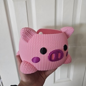 May include: A pink, pig-shaped container with a knitted texture. It features black eyes, a purple snout, and purple cheek spots. The ears and feet are also pink. The container is held in a hand.