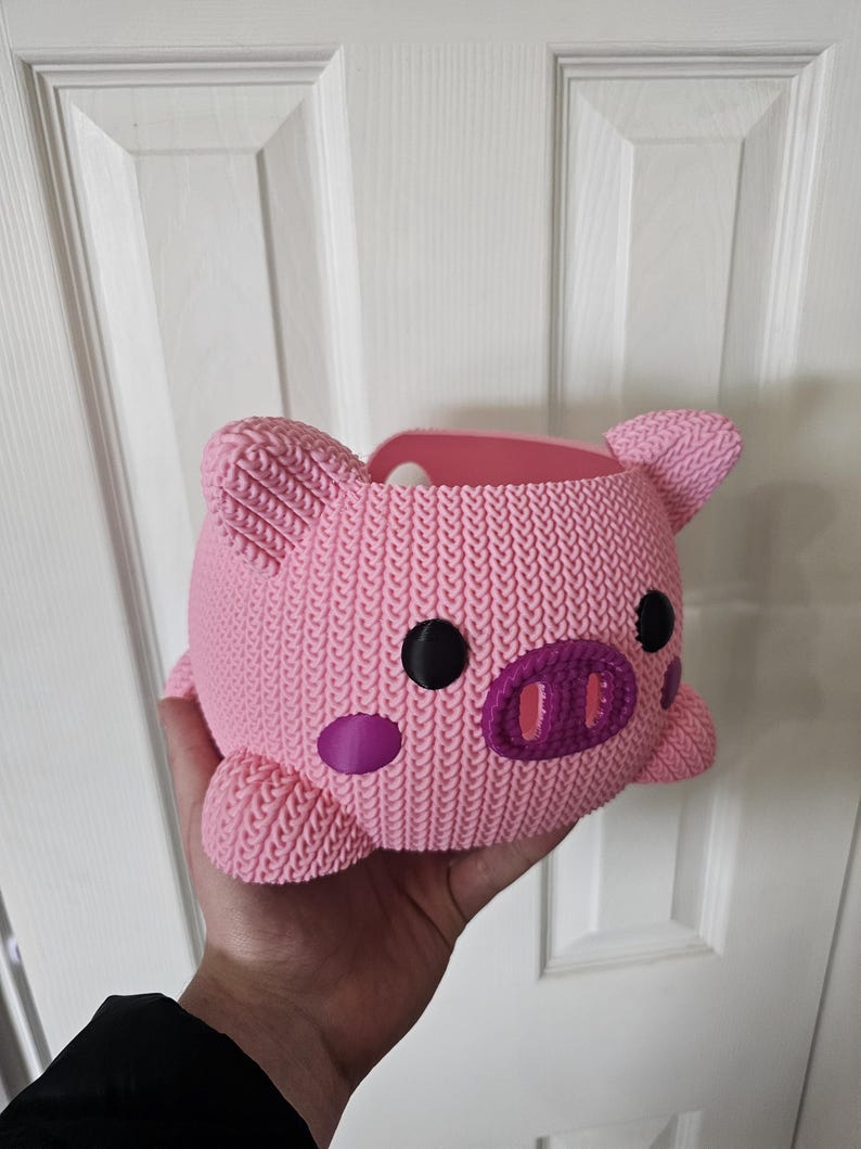 May include: A pink, pig-shaped container with a knitted texture. It features black eyes, a purple snout, and purple cheek spots. The ears and feet are also pink. The container is held in a hand.