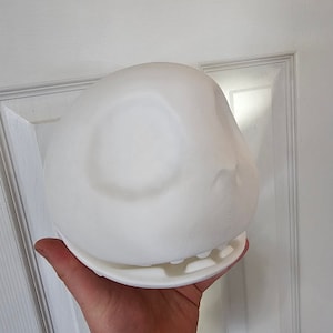 May include: A white, spherical object with a flat base and an open-mouth design. The object is held in a person's hand, showing its size and shape. The design features a smooth, rounded top with a subtle indentation, and a detailed lower section.