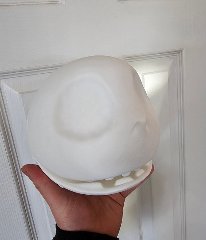 May include: A white, spherical object with a flat base and an open-mouth design. The object is held in a person's hand, showing its size and shape. The design features a smooth, rounded top with a subtle indentation, and a detailed lower section.