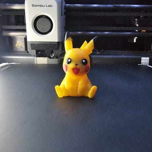 May include: A yellow Pikachu figure with red cheeks and a smiling face, sitting on a dark surface. The figure is 3D printed. A 3D printer with the "Bambu Lab" logo is in the background.
