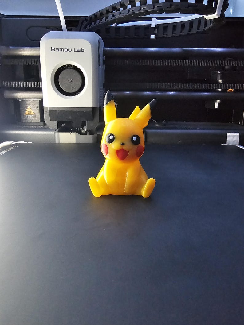 May include: A yellow Pikachu figure with red cheeks and a smiling face, sitting on a dark surface. The figure is 3D printed. A 3D printer with the "Bambu Lab" logo is in the background.