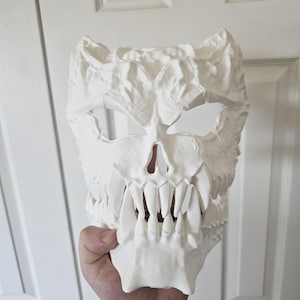 May include: A white mask with a skull design and a textured surface. The mask has sharp teeth, eye and nose openings, and a rough, uneven surface. The mask is held up by a hand.