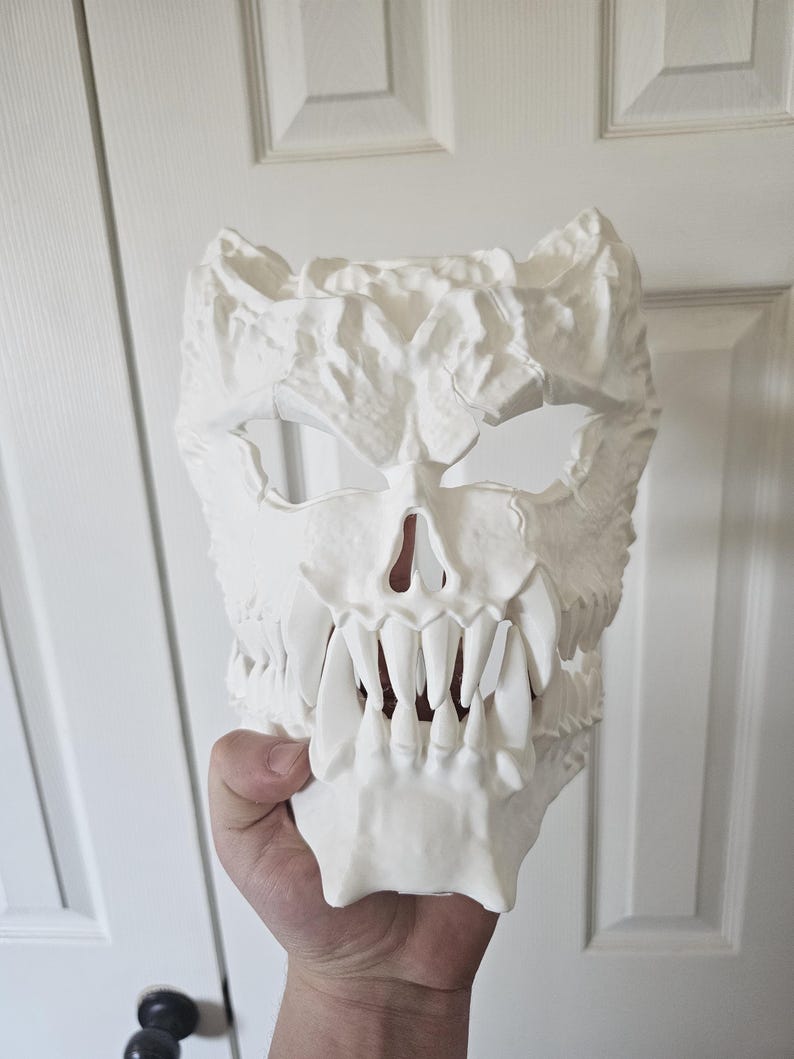 May include: A white mask with a skull design and a textured surface. The mask has sharp teeth, eye and nose openings, and a rough, uneven surface. The mask is held up by a hand.