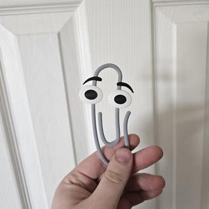 May include: A grey paperclip with cartoon eyes and eyebrows. The paperclip is held in a hand against a white door background. The eyes are white with black pupils and black eyebrows.