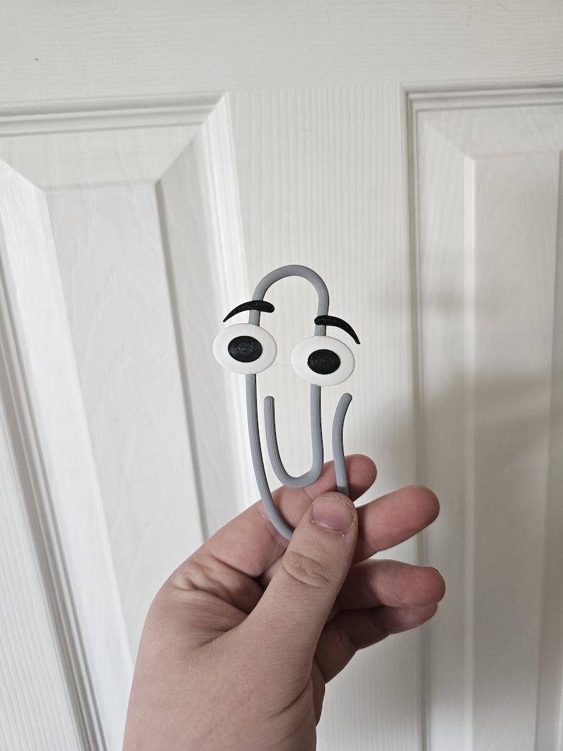 May include: A grey paperclip with cartoon eyes and eyebrows. The paperclip is held in a hand against a white door background. The eyes are white with black pupils and black eyebrows.
