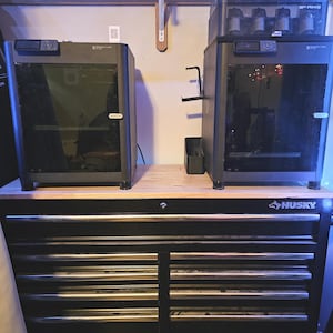 May include: Two dark gray 3D printers with glass doors sit atop a black Husky tool chest. The printers have a sleek, modern design with integrated cameras. The tool chest has multiple drawers with silver handles.