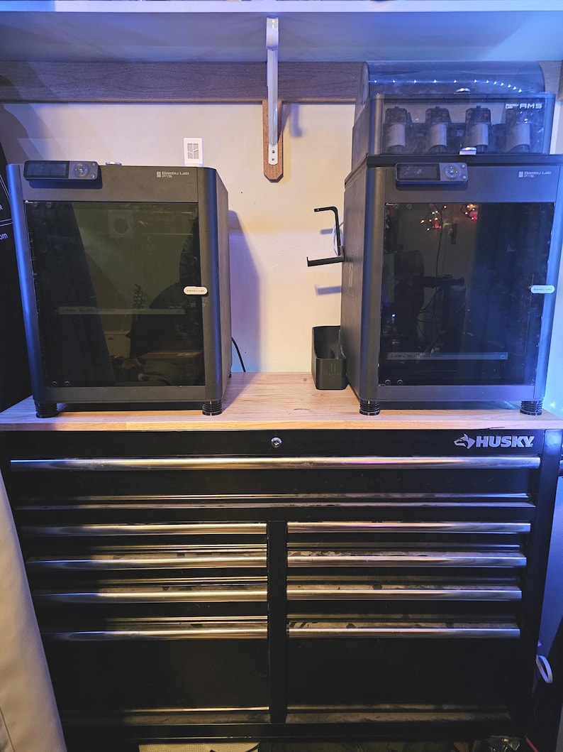 May include: Two dark grey 3D printers with glass doors are positioned on a black Husky tool chest. The printers have a modern design with cameras. The tool chest has multiple drawers with silver handles.