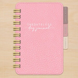 May include: A pink leather-bound journal with a gold spiral binding. The cover has the text "THE DATELESS day journal" in white lettering. The journal has three tabs on the right side labelled "QUICK LINKS", "NOTES", and "PLANNER".