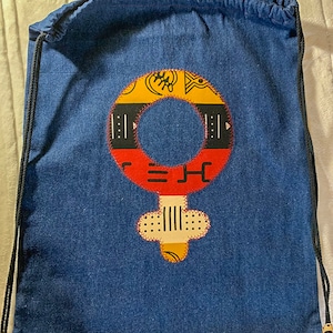 Può includere: Una borsa a cordoncino blu con un designo di simbolo femminile colorato. Il simbolo è realizzato con tessuti di diversi colori e presenta un motivo geometrico.