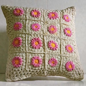 May include: Square crochet pillow with a beige background and pink and yellow flower accents. The flowers are evenly spaced across the pillow's surface, creating a floral pattern. The pillow is a square shape.