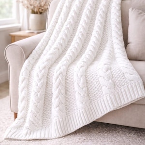 May include: A cream cable-knit throw blanket on a beige sofa. The blanket has a chunky, textured weave with bold cable patterns and a ribbed edge. The cosy blanket adds warmth and texture to the living space, perfect for snuggling.