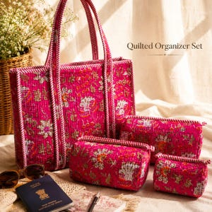 Op de afbeelding: Een fuchsia gekleurde, gewatteerde organizer set, bestaande uit een tote bag en drie kleinere pouches. Het heeft een gouden en witte bloemenpatroon en is afgewerkt met een witte en roze touwachtige rand. De tekst "Quilted Organizer Set" is zichtbaar.