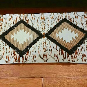 Puede incluir: Una alfombra rectangular de color blanquecino con un patrón repetitivo marrón y blanco. Tres grandes formas de diamante con un diseño de estrella blanca están centradas en la alfombra. Los diamantes están delineados en negro y marrón. La alfombra tiene flecos en ambos extremos.