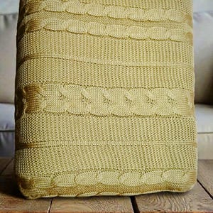 May include: A light yellow knitted throw blanket with a cable knit design. The blanket is folded and placed on a wooden table. The cable knit pattern is a prominent feature, adding texture and visual interest to the soft, cozy blanket.