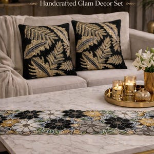 Peut inclure: Un ensemble de décoration de luxe pour la maison comprenant deux coussins noirs avec des motifs de feuilles dorées, un chemin de table perlé avec des motifs floraux et un plateau doré avec des bougies. Le texte indique "Luxury Black Gold Silver Beaded Bundle".