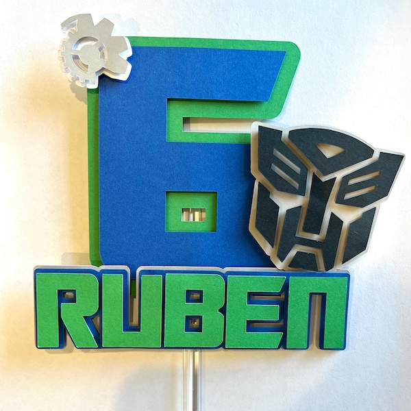 Könnte beinhalten: Ein grüner und blauer Tortendekor mit der Zahl 6, dem Wort "RUBEN", einem silbernen Zahnrad und einem schwarzen Transformers-Logo.