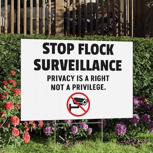 May include: A white rectangular yard sign with the text "STOP FLOCK SURVEILLANCE" and "PRIVACY IS A RIGHT NOT A PRIVILEGE." A red circle with a crossed-out security camera is at the bottom. The sign is on metal stakes in a grassy yard.
