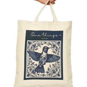 May include: A white canvas tote bag with blue handles. The bag features a blue and white illustration of a hummingbird in a floral frame. The text "oma things" is printed in cursive above the image.