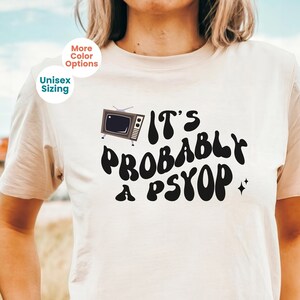 May include: Beige unisex t-shirt with the phrase "It's Probably a Psypop" in black, retro-style lettering. A small, vintage television graphic is on the left. The shirt also has "More Color Options" and "Unisex Sizing" text.