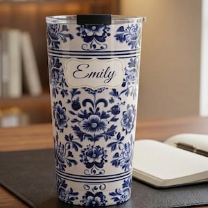 May include: A cylindrical tumbler with a white base and a blue floral pattern reminiscent of delftware. The centre features a decorative label with the name Emily printed in a dark blue script font. The tumbler has a black lid on top.