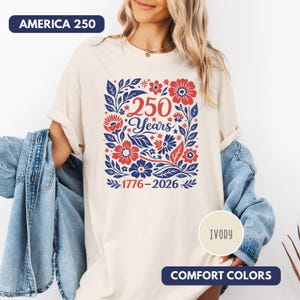 May include: Ivory-colored t-shirt with a floral design in red and blue, featuring the text "250 Years" and the dates "1776-2026". The shirt also has the text "AMERICA 250" and "COMFORT COLORS".