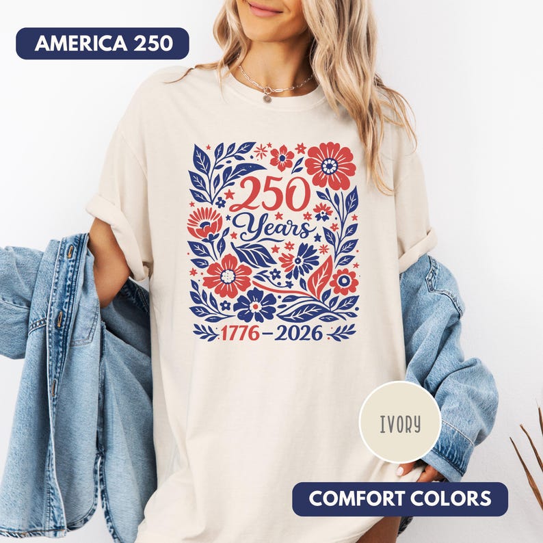 May include: Ivory-colored t-shirt with a floral design in red and blue, featuring the text "250 Years" and the dates "1776-2026". The shirt also has the text "AMERICA 250" and "COMFORT COLORS".