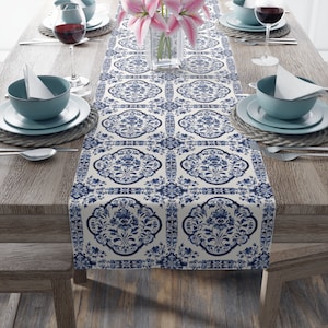 Puede incluir: Un camino de mesa blanco con un patrón floral azul y blanco. El camino de mesa está sobre una mesa de madera con cubiertos a cada lado.