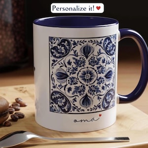 May include: White ceramic mug with a blue and white floral pattern. The mug has a blue rim and handle. The word "oma" is written on the bottom of the mug in cursive.