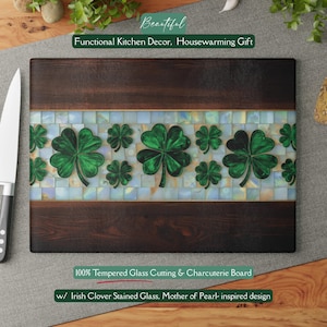 May include: A rectangular cutting board with a dark wood border and a central mosaic design. The mosaic features a pattern of green shamrocks on a mother-of-pearl inspired background. Text on the board reads "100% Tempered Glass Cutting & Charcuterie Board".