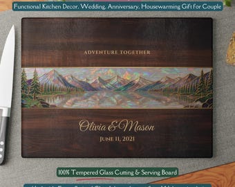 May include: A rectangular cutting board with a faux walnut trim design. The center features a lakeside forest stained glass inlay with mountains and trees. The text "ADVENTURE TOGETHER" is above the names "Olivia & Mason" and the date "JUNE 11, 2021".