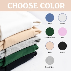 Puede incluir: Una pila de sudaderas plegadas en varios colores, incluyendo blanco, beige, gris, verde y negro. La imagen tambi&eacute;n muestra opciones de color: azul marino, blanco, verde bosque, rosa, arena, negro y gris deportivo. El texto dice "CHOOSE COLOR".