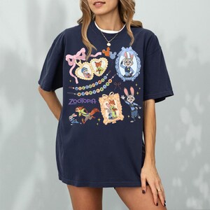 May include: Navy blue t-shirt with characters from the film Zootopia. The design includes framed images of Judy Hopps and Nick Wilde, and the word "Zootopia" in purple. The shirt has a relaxed fit.