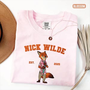 Puede incluir: Camiseta rosa claro con la inscripci&oacute;n bordada en naranja "NICK WILDE" sobre una ilustraci&oacute;n de un zorro con camisa estampada y pantal&oacute;n. El texto "EST. 2025" est&aacute; debajo. Tambi&eacute;n se ve un collar dorado y un sombrero.