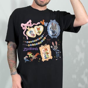 May include: Black t-shirt with cartoon images and the word "Zootopia". The design includes framed portraits of characters, a pink bow, and a string of beads. The shirt has a crew neck and short sleeves.