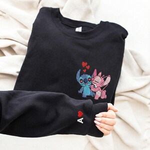 Pu&ograve; includere: Felpa nera con un ricamo di una coppia di cartoni animati blu e rosa con cuori rossi. La manica sinistra ha un cuore rosso e la lettera "A". La felpa &egrave; piegata su una superficie bianca.