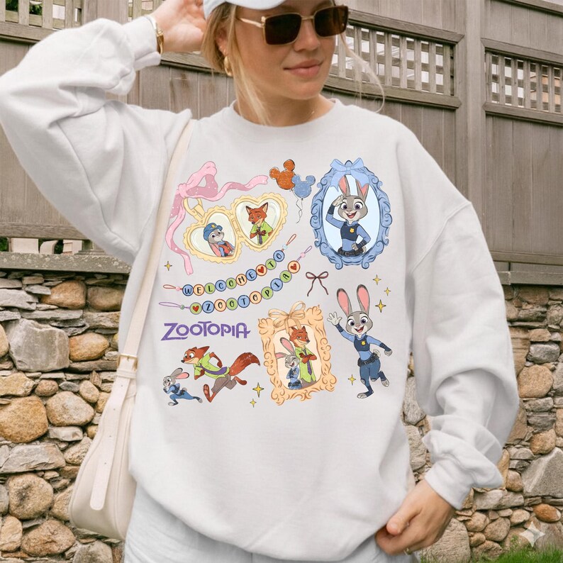 May include: White sweatshirt with colourful illustrations of characters and the word "Zootopia." The design includes framed images, hearts, and a banner with text. The sweatshirt has long sleeves and a crew neck.