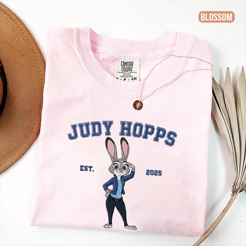 Puede incluir: Una camiseta rosa claro con la inscripci&oacute;n "JUDY HOPPS" en azul y una ilustraci&oacute;n de un conejo con una chaqueta azul. La camiseta tambi&eacute;n incluye el texto "EST. 2025". Tambi&eacute;n se ven un collar dorado y un sombrero marr&oacute;n.