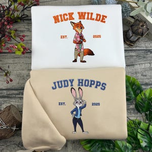 Puede incluir: Dos sudaderas, una blanca y otra beige, con personajes de dibujos animados bordados. La sudadera blanca muestra "NICK WILDE" sobre un personaje de zorro, y la beige muestra "JUDY HOPPS" sobre un personaje de conejo. Ambas incluyen "EST. 2025".