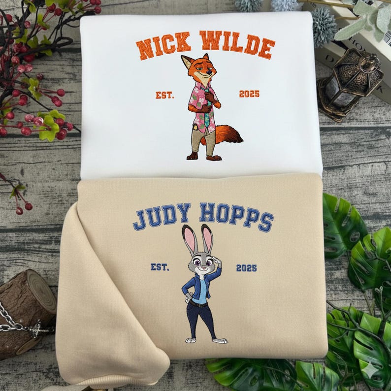 Puede incluir: Dos sudaderas, una blanca y otra beige, con personajes de dibujos animados bordados. La sudadera blanca muestra "NICK WILDE" sobre un personaje de zorro, y la beige muestra "JUDY HOPPS" sobre un personaje de conejo. Ambas incluyen "EST. 2025".
