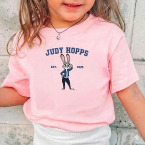 Puede incluir: Una camiseta rosa claro con la inscripci&oacute;n "JUDY HOPPS" en azul, encima de una imagen de dibujos animados de Judy Hopps de Zootopia. La imagen muestra a Judy con una chaqueta azul y pantalones oscuros. El texto "EST. 2025" tambi&eacute;n est&aacute; en la camiseta.