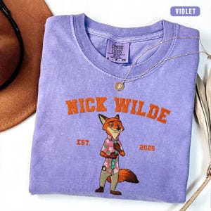 Puede incluir: Una camiseta violeta Comfort Colors con un dise&ntilde;o bordado de Nick Wilde. La camiseta tiene el texto "NICK WILDE EST. 2025" en naranja. Un collar dorado y un sombrero marr&oacute;n son visibles en el fondo.