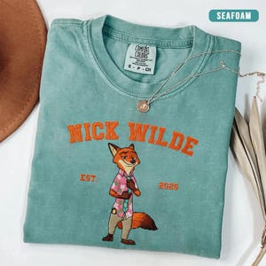Puede incluir: Camiseta verde mar con el texto bordado en naranja "NICK WILDE". La camiseta presenta un personaje de zorro de dibujos animados con una camisa rosa y pantalones color caqui. El texto "EST. 2025" tambi&eacute;n est&aacute; bordado en la camiseta.