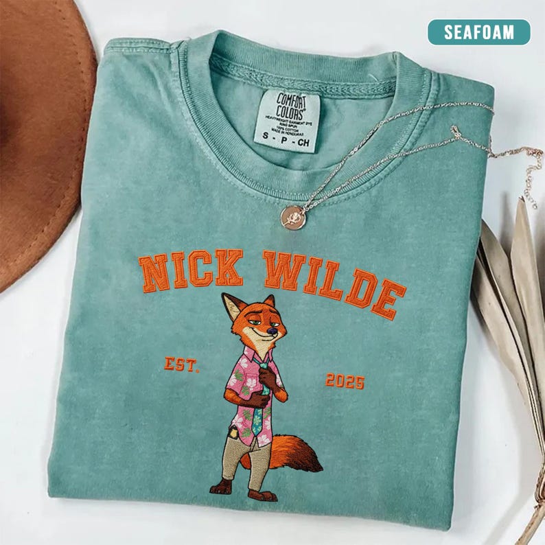 Puede incluir: Camiseta verde mar con el texto bordado en naranja "NICK WILDE". La camiseta presenta un personaje de zorro de dibujos animados con una camisa rosa y pantalones color caqui. El texto "EST. 2025" tambi&eacute;n est&aacute; bordado en la camiseta.
